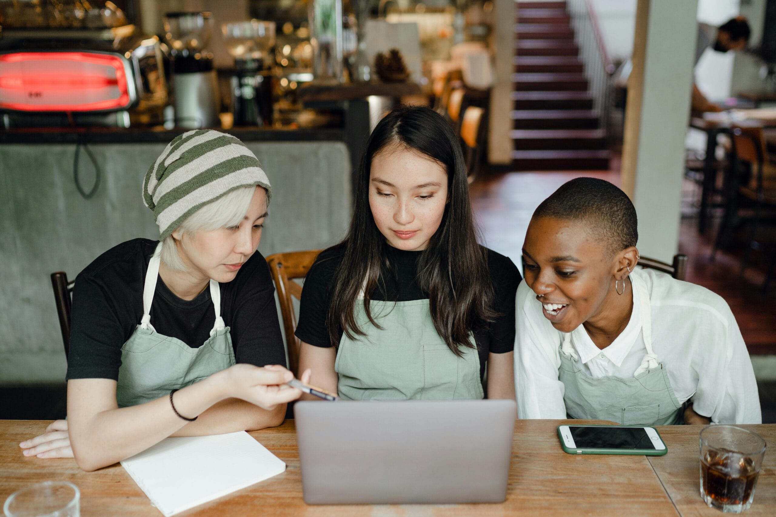 Three women in aprons collaborate at a table with a laptop, discussing strategies for their small businesses.