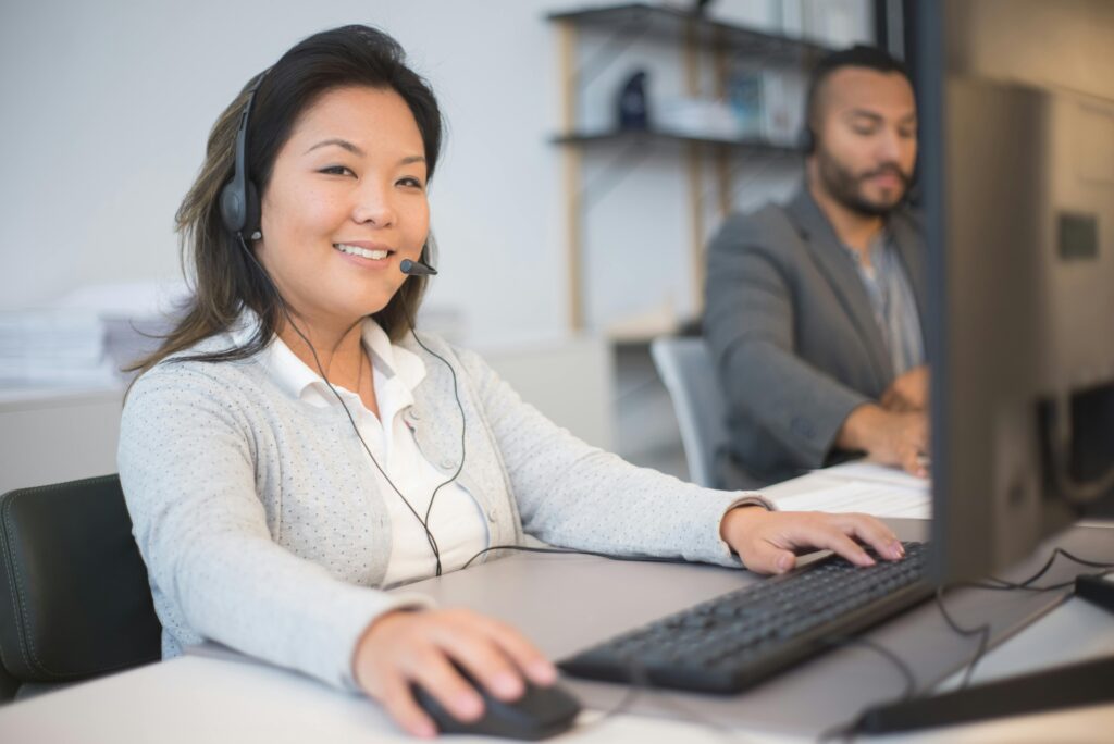 A woman with a headset smiles while working on a computer as a virtual receptionist for a call answering service.